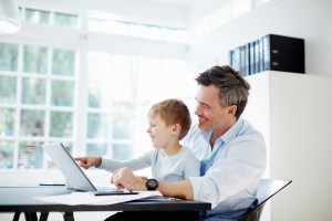 Man sitting at desk with son on his knee with laptop computer and paperwork - B2B